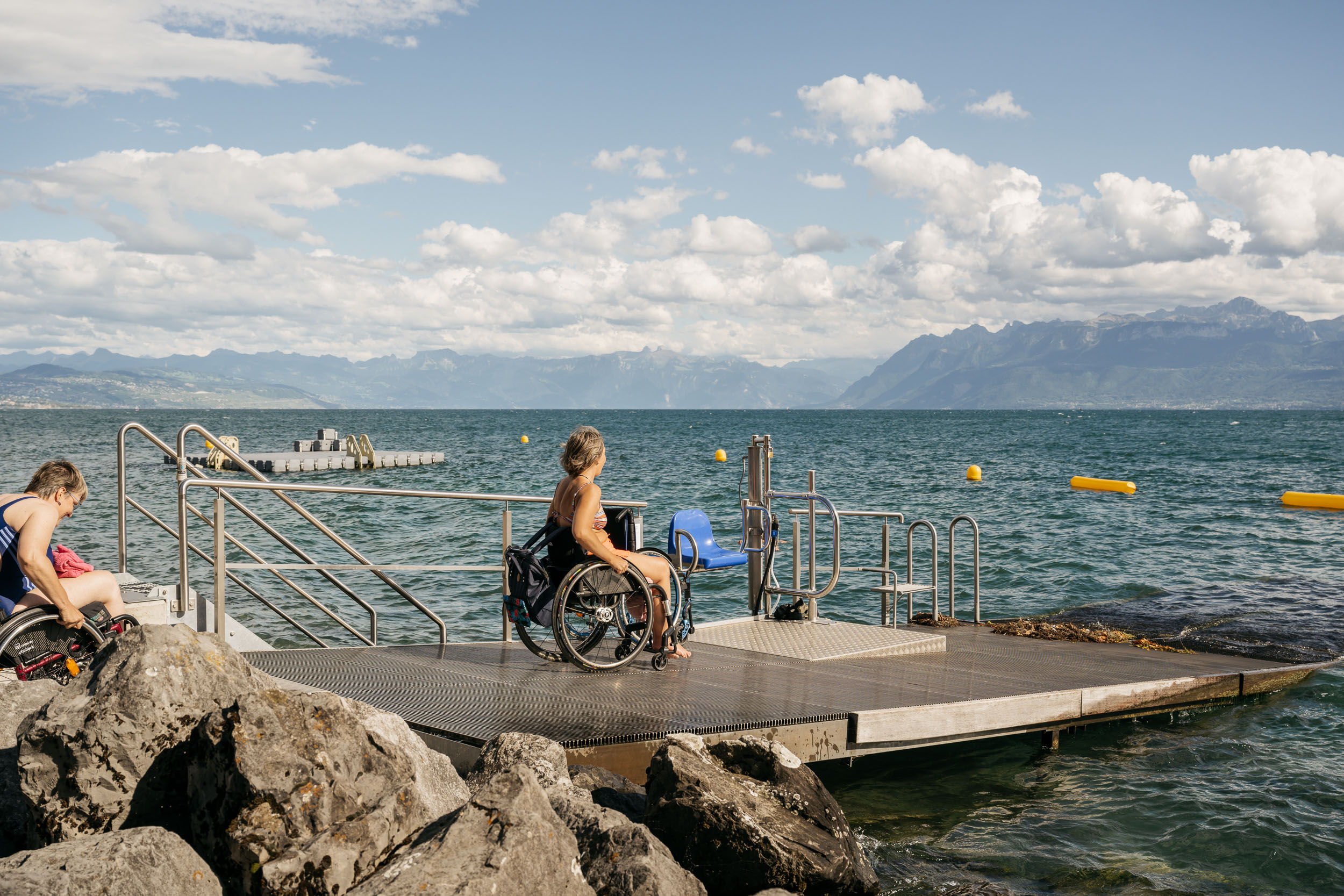Femme en chaise roulante, sur le ponton d'accès au lac. Monte-personne pour se mettre à l'eau. Lac et vue sur les montagnes.