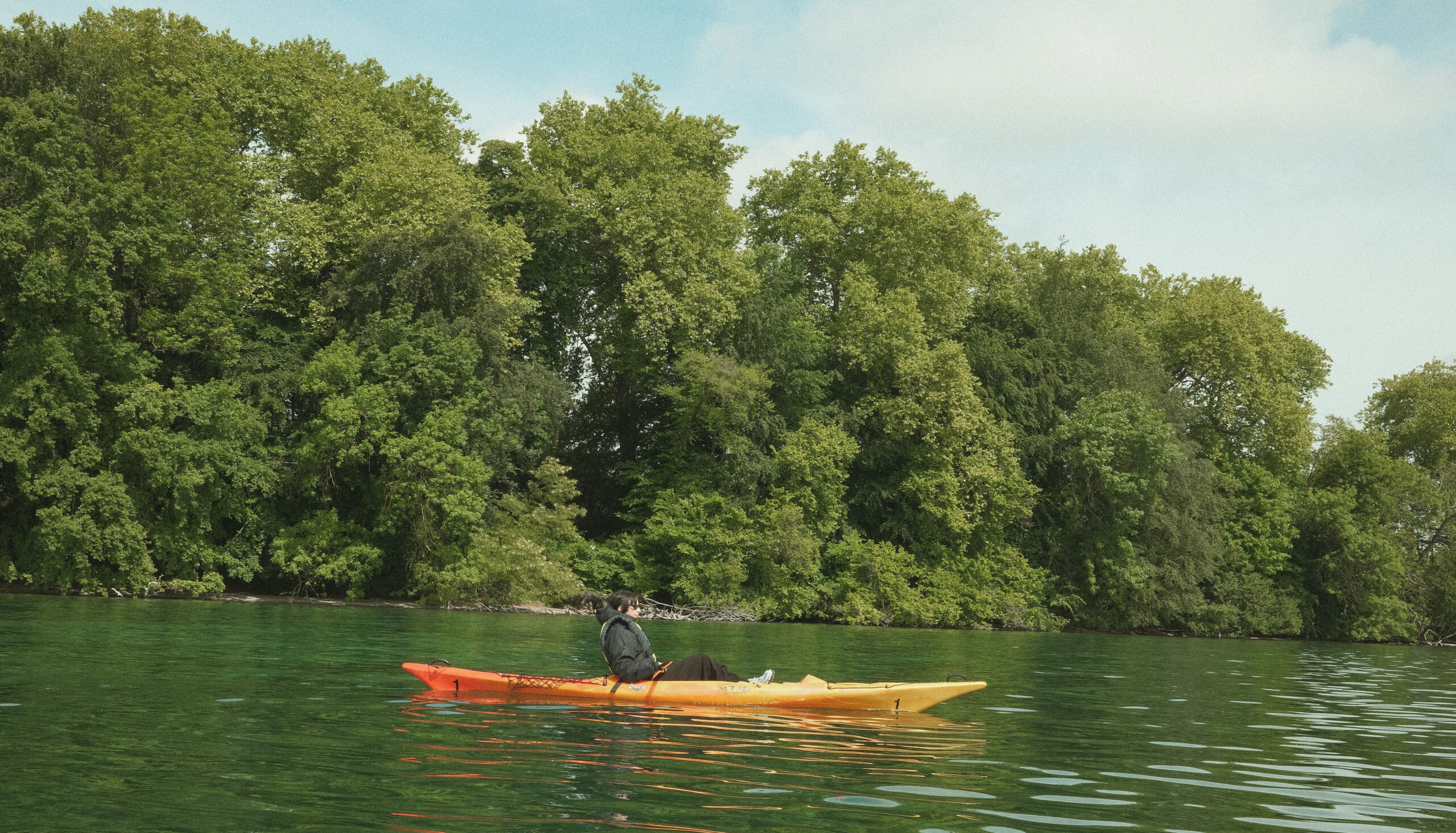 Une kayakiste assise dans un kayak orange et jaune navigue sur un lac aux eaux vertes, devant une rive bordée d’une forêt dense et verdoyante.