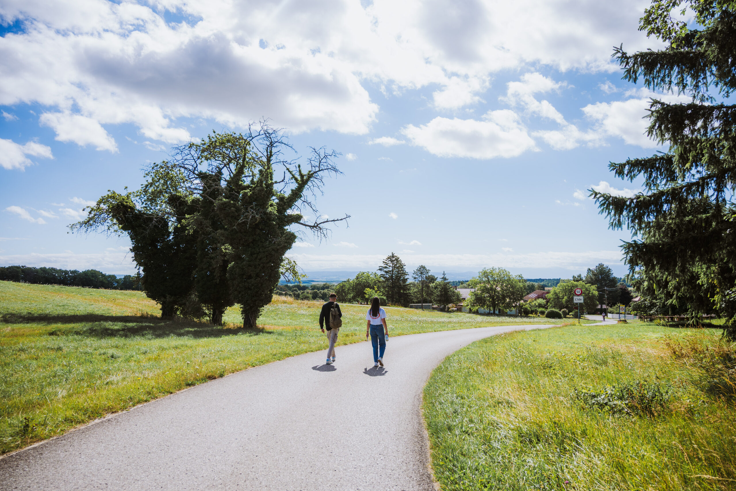 Deux personnes marchent sur une route de campagne entourée de prairies, avec des arbres, des maisons et un paysage vallonné à l’horizon.