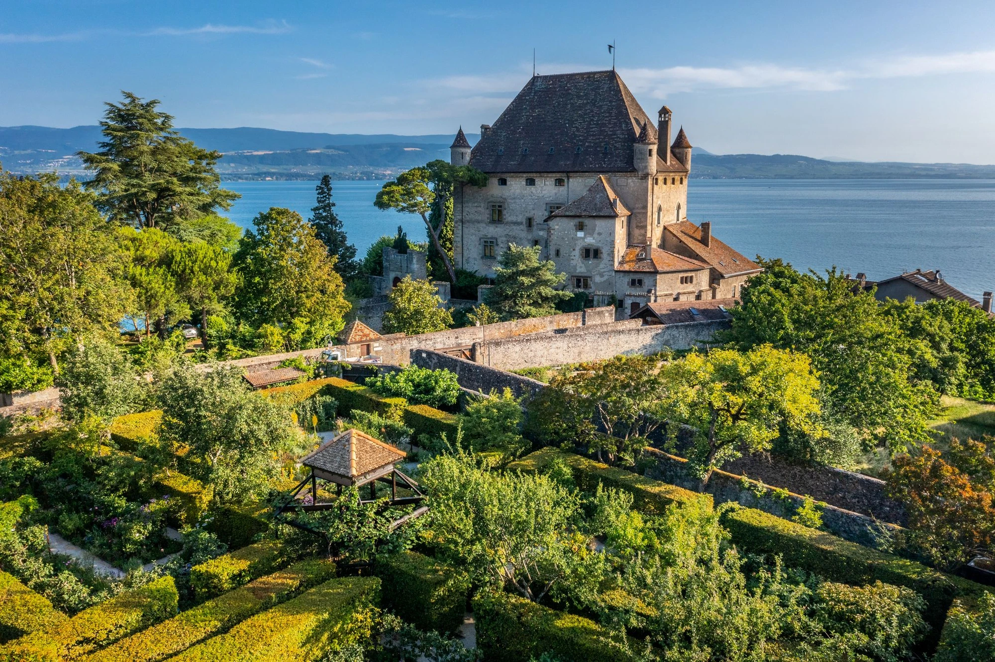 Vue aérienne du château d'Yvoire et de son jardin des cinq sens, avec haies taillées et un kiosque en bois, en bordure du lac Léman entouré de montagnes.