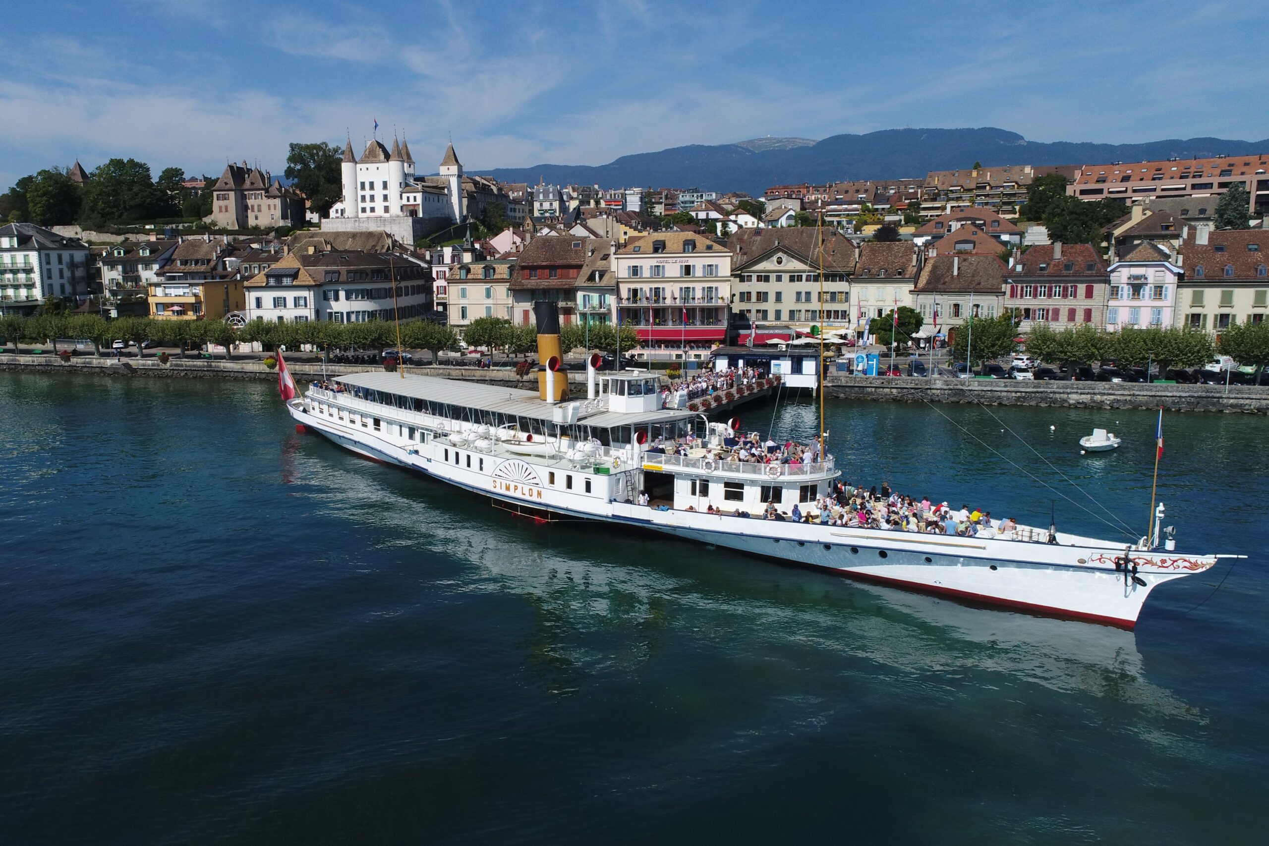 Vue aérienne du bateau à vapeur Simplon quittant le débarcadère de Nyon, avec le château de Nyon et les toits de la vieille ville en arrière-plan.