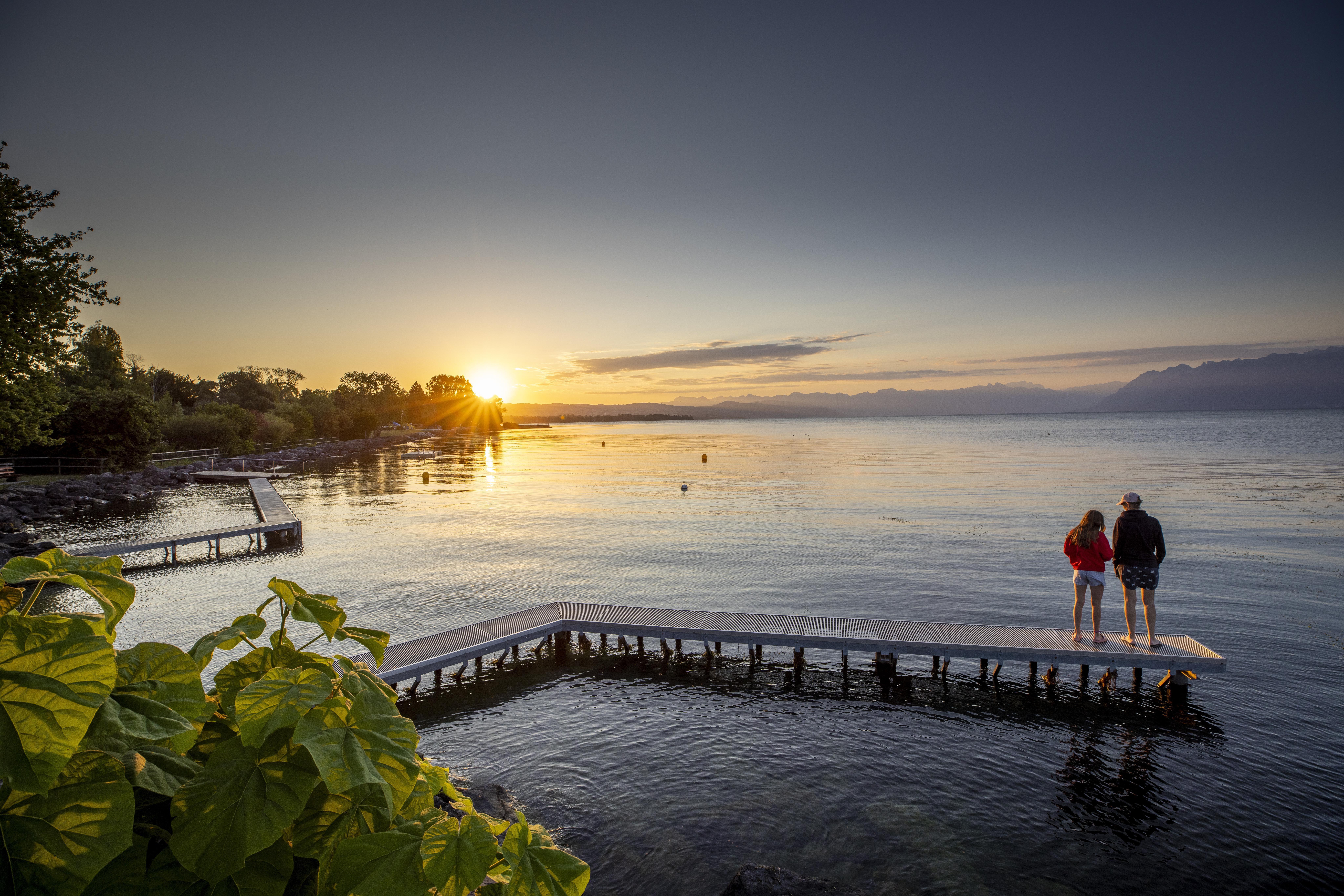 Coucher de soleil sur le lac Léman à Morges, avec un ponton au premier plan et deux personnes admirant la vue. Les montagnes se dessinent à l’horizon, tandis que des feuillages bordent la rive.